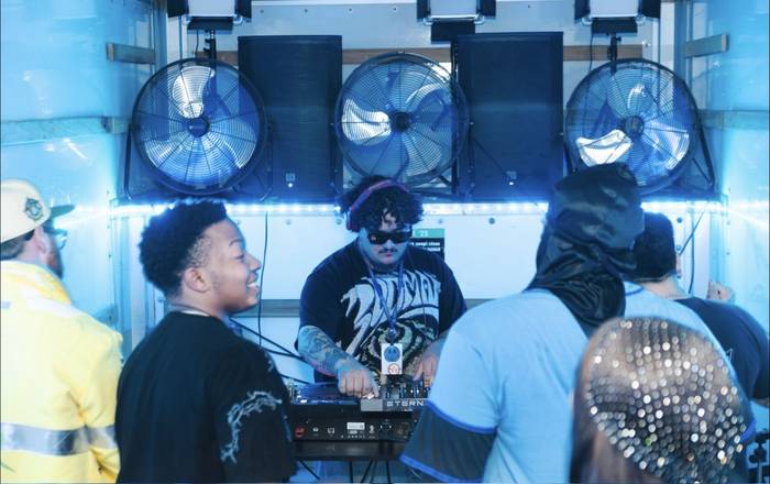 A DJ in sunglasses working the decks in a box truck lit hard blue, a friend laughing beside him